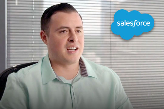 Man in green shirt speaking in an office with blue cloud Salesforce logo in top right corner.