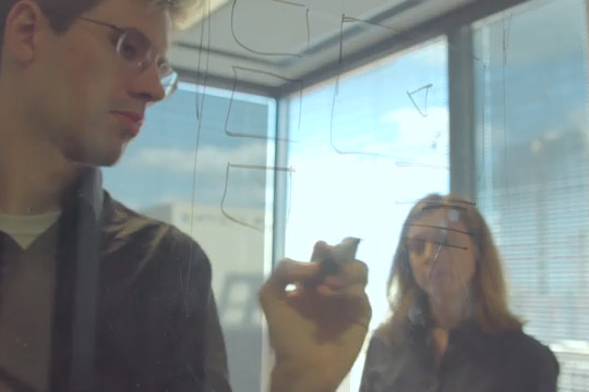 A woman watching a man draw on glass with a dry erase marker.