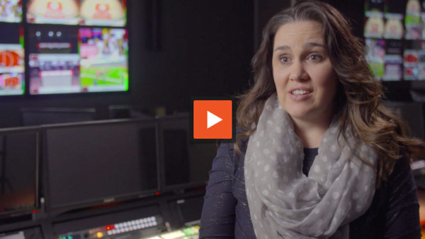 Woman in a gray polka-dot scarf speaking at a video production station, with orange play button over it.